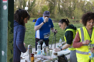 Moment de ravitaillement pour un homme qui fait de la marche nordique, il boit un gobelet d'eau que sert des bénévoles portant un gilet jaune, le stand est localisé au niveau du lac des Pêcheurs, on aperçoit le lac derrière eux - Agrandir l'image 15 sur 32, fenêtre modale