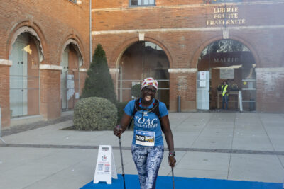 Une femme qui fait de la marche nordique fait un grand sourire alors qu'elle marche devant la Mairie - Agrandir l'image 19 sur 32, fenêtre modale