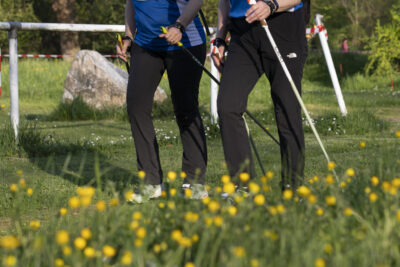 Deux femmes font de la marche nordique, sur un chemin d'herbe, à proximité d'une barrière blanche qui délimite un stade. Au premier plan on voit en flou les fleurs jaunes - Agrandir l'image 23 sur 32, fenêtre modale