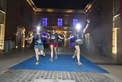 Trois femmes courent sur le parvis de la Mairie, en se tenant par la main, munies de lumières frontales, elles sont tout sourire et bras en l'air - Agrandir l'image 22 sur 32, fenêtre modale