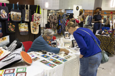 Une femme se penche au-dessus d'un stand pour observer les photos posées dessus. Derrière le stand; des tote bags avec dessins de fleurs et de peintures - Agrandir l'image 6 sur 17, fenêtre modale
