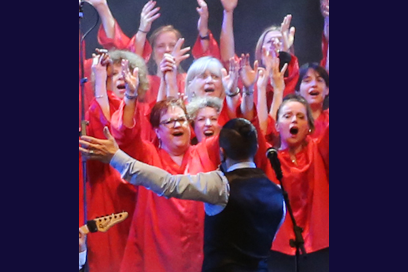 photo en plan rapproché du Choeur One Heart Gospel en toges rouges traditionnelles, bras levés sourires et bouches ouvertes en train de chanter avec au premier plan de dos le chef de choeur Loïc Geffray en train des les diriger en costume trois pièces - Agrandir l'image, fenêtre modale