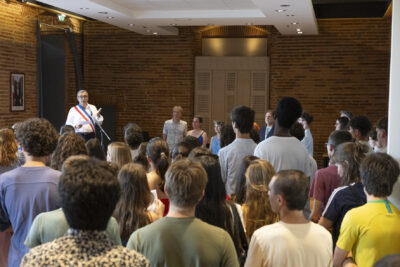 Le Maire fait un discours, avec l'écharpe tricolore, devant un groupe de jeunes réunis dans la salle du Conseil, qu'on voit de dos sur la photo. D'autres élus se tiennent debout à côté du Maire - Agrandir l'image 10 sur 10, fenêtre modale