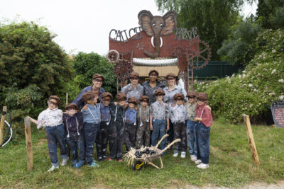 Photo d'un groupe d'enfants et animateurs déguisés avec jean, bretelle et chemise, bérets et masque sur les yeux, devant un stand couleur cuivre décoré à la main, une tête d'éléphant au sommet et écrit "cauch" et "rêve" - Agrandir l'image 24 sur 24, fenêtre modale
