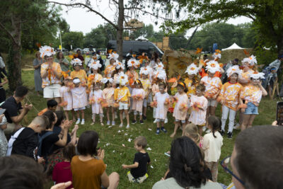 Photo d'un groupe d'enfants rassemblés avec leurs animateurs, habillés en orange et blanc et avec des marguerites en papier sur la tête - Agrandir l'image 14 sur 24, fenêtre modale
