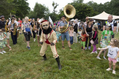Photo d'une personne en costume beige et rouge, avec un masque en papier mâché de créature, des enfants regardent et des musiciens la suivent - Agrandir l'image 12 sur 24, fenêtre modale