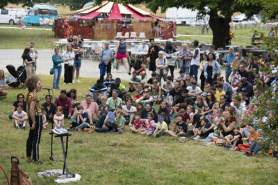 Photo paysage d'un spectacle en plein air, avec un public nombreux et familial assis dans l'herbe - Agrandir l'image 3 sur 24, fenêtre modale