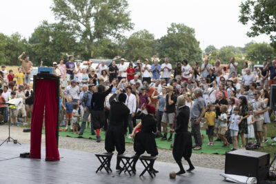 Photo paysage du spectacle sur la scène, le public est debout et imite les gestes de la troupe sur la scène - Agrandir l'image 10 sur 24, fenêtre modale