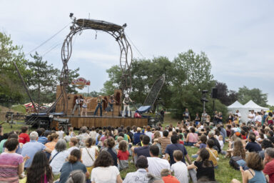 Photo paysage d'un spectacle d'acrobatie, une troupe en costume de cowboy est sur une scène surmontée, le public familial et nombreux assis devant dans l'herbe