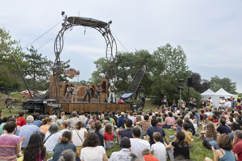 Photo paysage d'un spectacle d'acrobatie, une troupe en costume de cowboy est sur une scène surmontée, le public familial et nombreux assis devant dans l'herbe