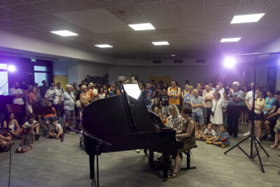 Photo d'une salle dans laquelle deux femmes jouent au piano, et de nombreuses personnes, dont des enfants assis devant, écoutent le concert - Agrandir l'image 7 sur 16, fenêtre modale