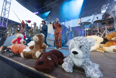 Photo d'une scène, un groupe se produit, des hommes vêtus de costumes façon pyjama, au premier plan le focus est fait sur des peluches - Agrandir l'image 8 sur 16, fenêtre modale