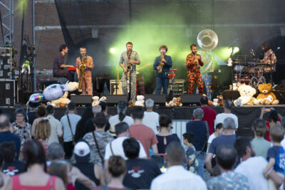 Photo paysage de la scène Place de la Mairie prise depuis le public, un groupe se produit, des hommes vêtus de costumes façon pyjama, avec des ours en peluche rassemblés sur le devant de la scène - Agrandir l'image 9 sur 16, fenêtre modale
