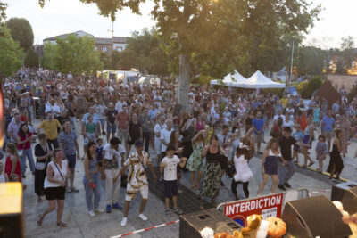 Photo du public nombreux rassemblé Place de la Mairie, devant la scène les gens dansent, plus loin on aperçoit les food trucks et des gens assis pour manger - Agrandir l'image 3 sur 16, fenêtre modale