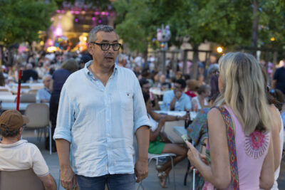 Photo paysage du Maire Frédéric Parre qui échange avec une femme blonde Place de la Mairie, on voit derrière eux les tables longues où des gens sont installés pour manger - Agrandir l'image 2 sur 16, fenêtre modale