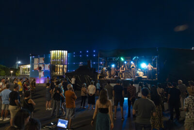 Photo paysage de nuit de la scène devant l'Escale qu'on voit en arrière plan, un groupe se produit tandis que des personnes assistent au concert - Agrandir l'image 11 sur 16, fenêtre modale