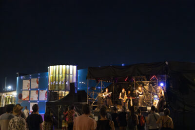 Photo paysage de nuit de la scène devant l'Escale qu'on voit en arrière plan, un groupe se produit tandis que des personnes assistent au concert - Agrandir l'image 5 sur 16, fenêtre modale