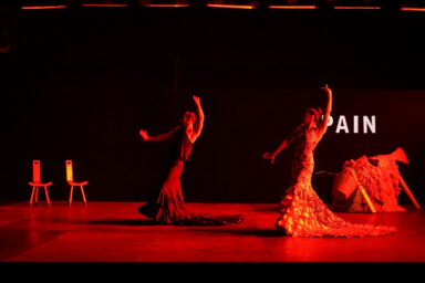 eux danseurs de flamenco en pleine représentation sur une scène faiblement éclairée par un éclairage rouge intense. Leurs postures intenses et leurs bras tendus expriment l'émotion et la passion caractéristiques du flamenco. Tous deux sont vêtus de longues robes traditionnelles à volants. En arrière-plan, le mot « ESPAGNE » apparaît en partie, évoquant le contexte culturel du spectacle. D'un côté de la scène, deux simples chaises en bois sont vides, et de l'autre, une pile de costumes ou d'accessoires repose au sol, renforçant l'atmosphère théâtrale. La scène est évocatrice, mettant en valeur le drame et l'expressivité du mouvement.