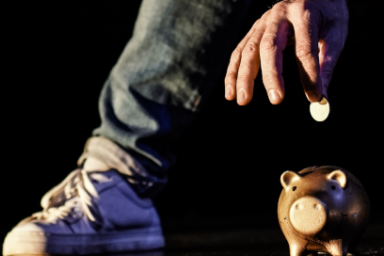 Une personne dont on n'aperçoit que la main et le pied en jean basket met une pièce dans une tirelire en forme de cochon posée au sol.