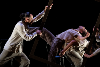 L'image montre une scène de danse contemporaine sur fond noir. On distingue plusieurs danseurs, dont un au premier plan à gauche tenant un cadre en bois. Les autres danseurs semblent interagir entre eux, certains étant assis sur des chaises ou en appui les uns sur les autres. L'éclairage met en valeur les mouvements et les expressions des danseurs.