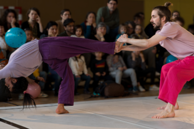 Une personne effectue une performance de danse aérienne dans une salle de spectacles, devant un public. La composition de l'image met en avant une personne au centre, en train de se déplacer dans les airs. La personne en vol est au centre du cadre. L'arrière-plan est composé d'un grand nombre de spectateurs, dont les visages et les corps sont flous, mais présents. L'action se déroule dans une salle d'un gymnase ou d'un auditorium.