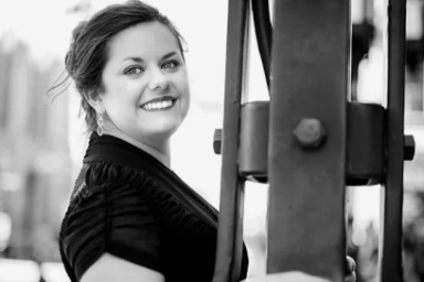 photo en noir et blanc de la soprano Anaïs Constans debout dans la rue dans un décor digne des temps modernes de Charlie Chaplin, en robe noire élégante et bras nus, elle tourne sa tête de trois quarts en direction de l'objectif, regard et sourire brillants, longs cheveux bruns attachés en arrière.