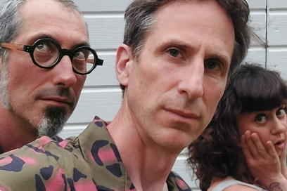 photo en plan rapproché pris contre la façade d'un garage du trio qui forme le groupe Jules Nectar le chanteur au premier plan en chemise hawaïenne, les deux musiciens, homme grisonnant à lunettes rondes montures noires et barbichette et femm, brunette malicieuse à la chevelure ondulée avec frange, qui l'accompagnent en arrière-plan, la mine faussement surprise et souriante. - Agrandir l'image, fenêtre modale