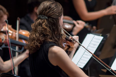 Plan rapproché sur une jeune violoniste cheveux attachés ondulés et brillants, de dos, au milieu des autres musiciens assis en train de jouer pendant un concert - Agrandir l'image, fenêtre modale