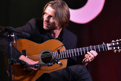 Le musicien guitariste, sur scène, assis sur un fauteuil cosy de profil, durant un concert, cheveux mi-longs, regard bleu tourné vers la gauche et souriant. - Agrandir l'image, fenêtre modale
