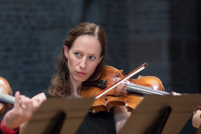 La violoniste, visage émergeant par-dessus les pupitres, son instrument au vernis brillant appuyé tendrement sur son épaule gauche, joue en plan rapprochée, traits concentrés et regard partant vers la gauche en direction du directeur musical, au milieu des autres violonistes que l'on devine. - Agrandir l'image, fenêtre modale