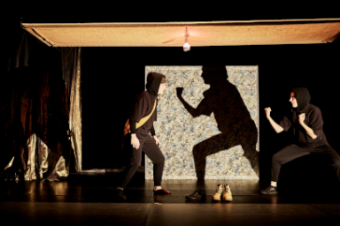 Une scène théâtrale met en scène deux personnages, en noir, en position de combat, avec des ombres projetées sur un mur. La composition de la scène est centrée sur l'interaction entre deux personnages. Les deux figures sont placées de part et d'autre d'un grand tableau, leurs silhouettes formant un contraste marqué avec la texture et le motif du tableau. Les personnages semblent engagés dans un échange dynamique, leurs postures suggérant une confrontation physique imminente ou un exercice martial. L'éclairage est très noir, ce qui met en évidence les silhouettes et les contrastes. Le cadre est précis et ne laisse aucun doute sur le focus de la scène qui est sur l'action. Les personnages principaux sont deux individus habillés de vêtements noirs, vêtus d'une tenue athlétique. Ils prennent une posture de combat, avec leurs bras et jambes pliés, leurs poings levés. Les expressions faciales ne sont pas visibles, les cagoules dissimulant leur visage, mais leurs postures et leur position suggèrent une certaine détermination ou un état d'esprit concentré.