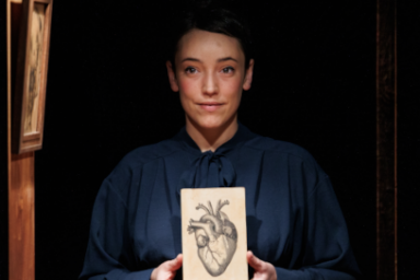 ur la scène plongée dans la pénombre, en plan rapproché, une comédienne du spectacle Entrañas de la compagnie El Patio Teatro tient entre ses mains le dessin au croquis d'un coeur se tient en robe d'époque de paysanne assise face au public à une table, son visage sage éclairé par la lumière