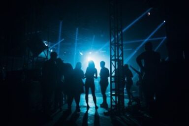photo en ombres chinoises de silhouettes debout aux abords d'une scène de concert dans la pénombre, éclairée par de puissants projecteurs bleus