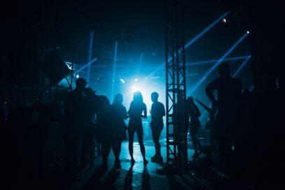 photo en ombres chinoises de silhouettes debout aux abords d'une scène de concert dans la pénombre, éclairée par de puissants projecteurs bleus - Agrandir l'image, fenêtre modale