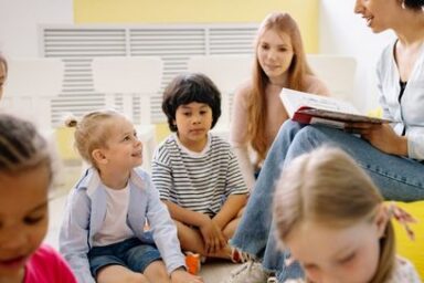Enfants assis en rond par terre autour des genoux d'une lectrice (à droite de l'image).