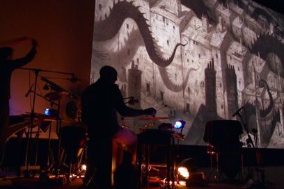 photo de ciné-concert vu depuis le bord de scène, des musiciens dans la pénombre utilisent leurs instruments et claviers électroniques sous la lumière de petites lampes de bureau tandis que le fond de scène affiche un écran géant où apparaissent des illustrations en théâtre d'ombres de tentacules surplombant un paysage urbain en sépia - Agrandir l'image, fenêtre modale