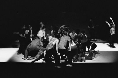 Photo d'ensemble, esthétique en mi-ombre, mi-lumière, en noir et blanc, du groupe des danseurs en tenue confortable en train d'effectuer une chorégraphie de groupe sur scène - Agrandir l'image, fenêtre modale