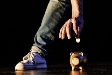 plan rapproché sur le bas du corps d'un comédien debout en train d emettre une pièce dorée dans une tire-lire en forme de porcelet sur scène entre ses pieds dans la lumière de scène qui éclaire sa jambe chaussée de basket