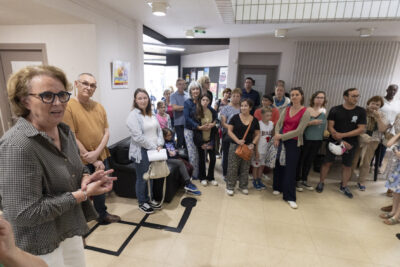 Au premier plan, Murielle Thomas l'adjointe au Maire déléguée à la culture se tient debout mains jointes s'adressant au public tandis qu'à ses côtés se tiennent les professeurs en arts visuels et dans l'entrée de l'Ecole les parents des élèves mineurs ainsi que les élèves adultes rassemblés. - Agrandir l'image 4 sur 7, fenêtre modale