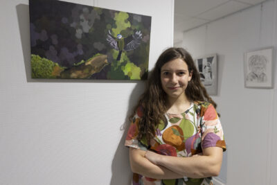 Accolée au pan de mur où est fixé un tableau de peinture aux couleurs vertes et grises qui évoquent un sous-bois, une jeune femme aux cheveux longs châtains, bras croisés, sourit à la caméra - Agrandir l'image 6 sur 7, fenêtre modale