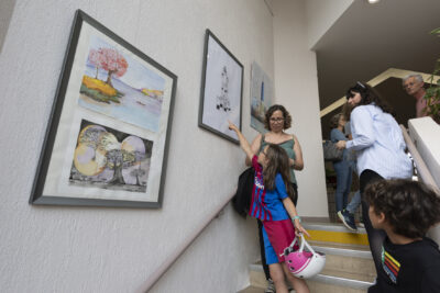 Dans le large escalier qui relie les étages de l'EEA, une jeune femme en robe décolletée couleur kaki, et sa fillette aux vêtements bariolés, tenant un casque de vélo rose, stationnent devant une série de grands tableaux qui mettent en valeur les aquarelles réalisées par des élèves en arts visuels. - Agrandir l'image 2 sur 7, fenêtre modale