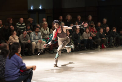 Dans un mouvement engagé debout en équilibre sur le pied gauche, le danseur en rollers est vêtu de manière fantaisiste d'un casque de protection de joueur de rugby, d'une cravate rouge volante autour de son cou nu, d'un débardeur de basketteur en filet noir; dans la main droite il tient une tringle à rideau de douche aux embouts ventousés qui fait office de baguette de majorette. Face à lui et en arrière-plan, le public est assis autour de la piste de rollers dans la salle du Phare de la Ville de Tournefeuille - Agrandir l'image 4 sur 14, fenêtre modale
