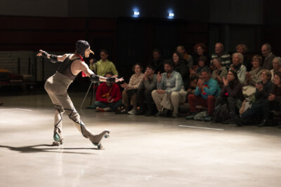 De profil, sur la piste éclairée, faisant apparaître un faux nez exagérément long, un danseur chaussés de rollers, en pantalon de ski et débardeur muni de protections anti-chute, fait une révérence au public assemblé autour de la piste - Agrandir l'image 1 sur 14, fenêtre modale