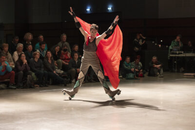 cape tenue dans son dos entre ses mains, le danseur à rollers effectue un freinage arrêt sur les talons, son corps figurant un grand X - Agrandir l'image 11 sur 14, fenêtre modale