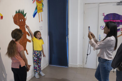 Une fillette en tee-shirt jaune citron debout contre le mur d'exposition,doigt tendu vers l'effigie d'un grand monstre velu à dents pointues au-dessus d'elle, pose devant sa mère de profil à droite de l'image, smartphone entre les mains - Agrandir l'image 3 sur 13, fenêtre modale