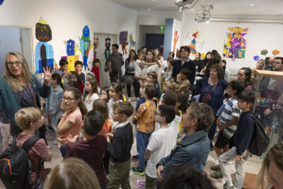 En salle d'exposition de la Médiathèque, le public nombreux écoute la'rtiste debout à gauche de l'image en train de présenter l'exposition des monstres colorés dessinés par les enfants, que l'on voit habiller les murs de la salle - Agrandir l'image 7 sur 13, fenêtre modale