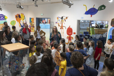 En salle d'exposition de la Médiathèque, le public nombreux écoute la'rtiste debout au centre de l'image, en train de présenter l'exposition des monstres colorés dessinés par les enfants, que l'on voit habiller les murs de la salle - Agrandir l'image 8 sur 13, fenêtre modale