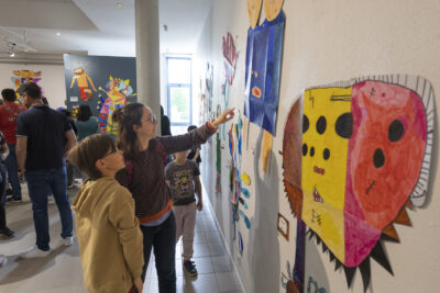 Une mère de bout à gauche, de profil, indique un élément de l'exposition du doigt sur le mur face à elle sous le regard appréciateur de son fils - Agrandir l'image 10 sur 13, fenêtre modale
