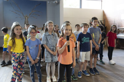 Un groupe d'enfants en tenues colorées se tient debout sur l'estrade du rez-de-jardin de la Médiathèque; au centre une fillette blonde aux traits malicieux présente l'exposition au micro face au public. - Agrandir l'image 1 sur 13, fenêtre modale