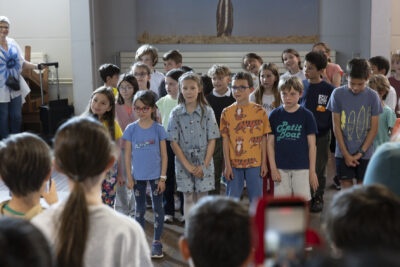 Les enfants rassemblés sur la petite scène du rez-de-jardin de la médiathèque chantonnent et dodelinent de la tête en souriant face au public dont on aperçoit les têtes au premier plan. - Agrandir l'image 2 sur 13, fenêtre modale